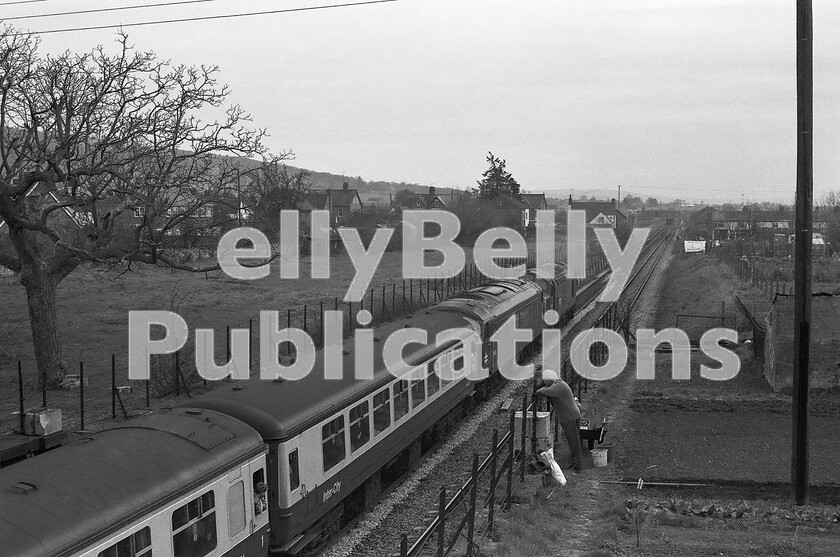 LPPC DSL BW 1193 
 Passengers in this southbound North and West express have just spent a Sunday lunchtime becoming very familiar with the location of the long-closed Bredon station, about two miles north of Ashchurch on the Birmingham-Bristol main line, after their Peak Class 45 failed. Help has arrived in the shape of an English Electric Class 37, presumably from the Gloucester direction on the engineers train seen in the far background. Rail ground staff are waiting for the train to cross to the northbound line to pass it by running wrong line. Meanwhile, the theatricals are being enjoyed by a local taking a break from his allotment. The hill on the left takes its name from the village and is Bredon Hill, after which A E Housman wrote his poem On Bredon Hill, after he finished A Shropshire Lad. 
 Keywords: BR, Bredon, Ashchurch, Class 37, Class 45, Drag, rescue, Failed, Passenger