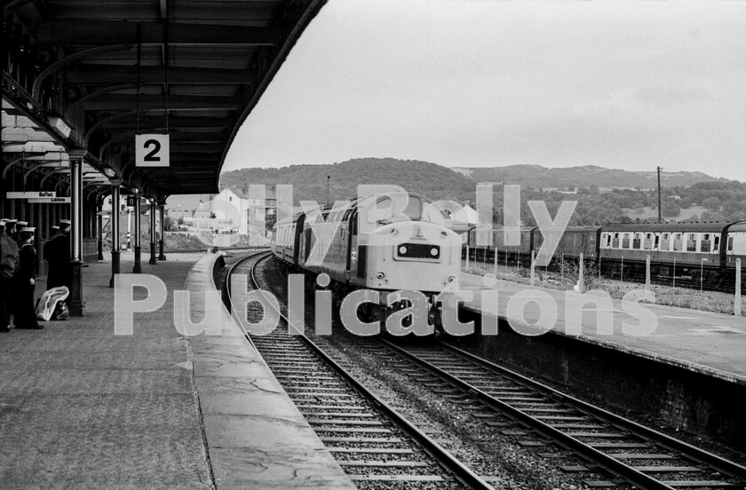 LPPC DSL BW 0779 
 Creeping through Llandudno Junction station so as not to stop before the signal ahead clears is one of the later English Electric Type 4 Class 40s. Numbers D345 to D399 were fitted with then-current fashion of central headcode-boxes when built and here is an immaculate, recently outshopped LMR example on its way to Holyhead with the daytime Irish Mail, timetabled to run non-stop from Chester to its destination. Its being watched by a group of sailors/ratings on the platform, presumably going home on leave, whilst on the right the ECS sidings seem to be occupied by stored BR GUVs, CCTs and Mark 1 coaches.