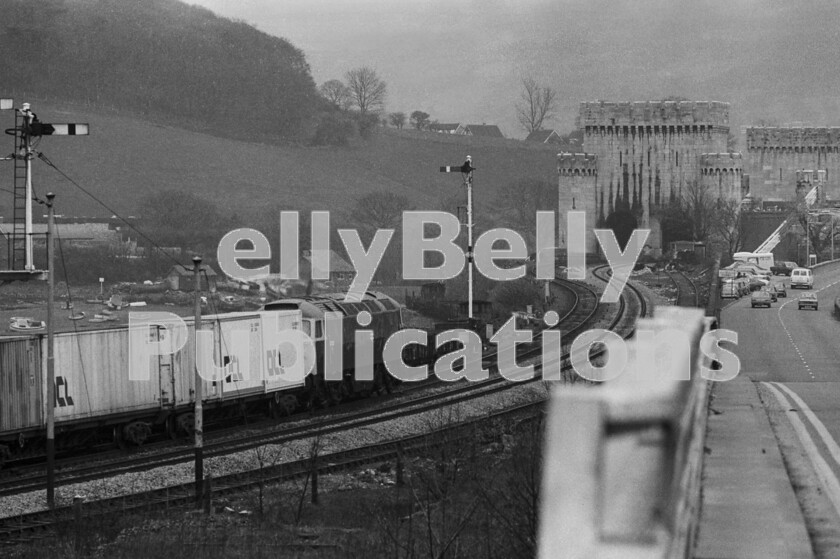 LPPC DSL BW 1163 
 Interlude at Llandudno Junction 4 The Freightliner has slowed to a stop at the signal which is set against it on the approach to Conway Castle bridge. Although the Britannia Bridge over the Menai Straits has always been famous for its method of conveying the railway in tubes, as can be seen, the Conway bridge was constructed on the same principle and is the only survivor as the former was destroyed by fire being rebuilt as a double-deck bridge, the railway sharing with the motorway standard A5 road which has also replaced the previous single-carriageway as seen here with a Ford Escort Mk2 and a Humber Sceptre amongst other 1970s vehicles. 
 Keywords: Digital, ISO, John Stiles